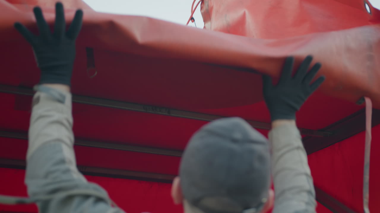 Back view of worker in uniform raising red truck tarp cover with both gloved hands during cargo preparation under daylight, revealing vehicle frame structure inside transport truck compartment