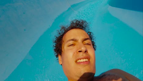 vista de la cara y el pecho de un hombre peludo con el pelo rizado deslizándose por un tobogán de agua azul a cámara lenta