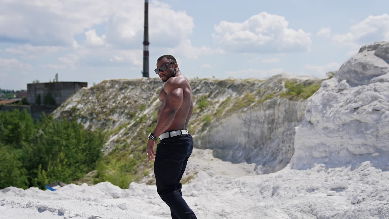 Athletic african man without shirt walks on hill. Muscular dark skinned sportsman among nature in summer. Slow motion.