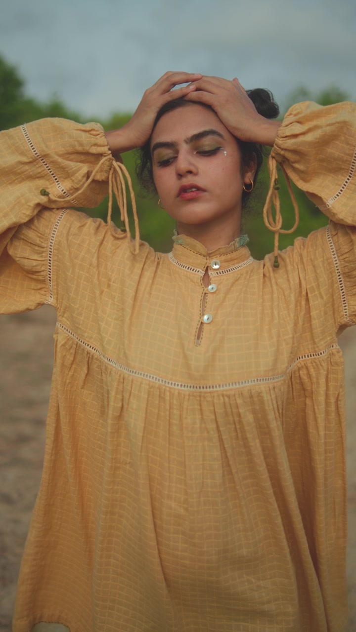 Young woman in yellow dress stands outdoors with eyes closed, hands on head, serene mood