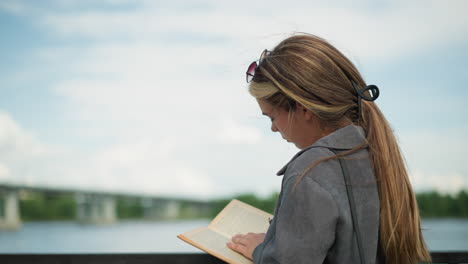 lady gazing into the distance focus back on her book while flipping to a new page of her book, resting her hand on an iron fence, the background features blurred greenery and a bridge over the river