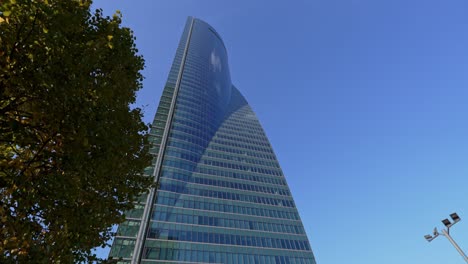 tilt down view of tall glass skyscrapper buildings seen through trees leaves against blue sky in madrid ctba bussines area