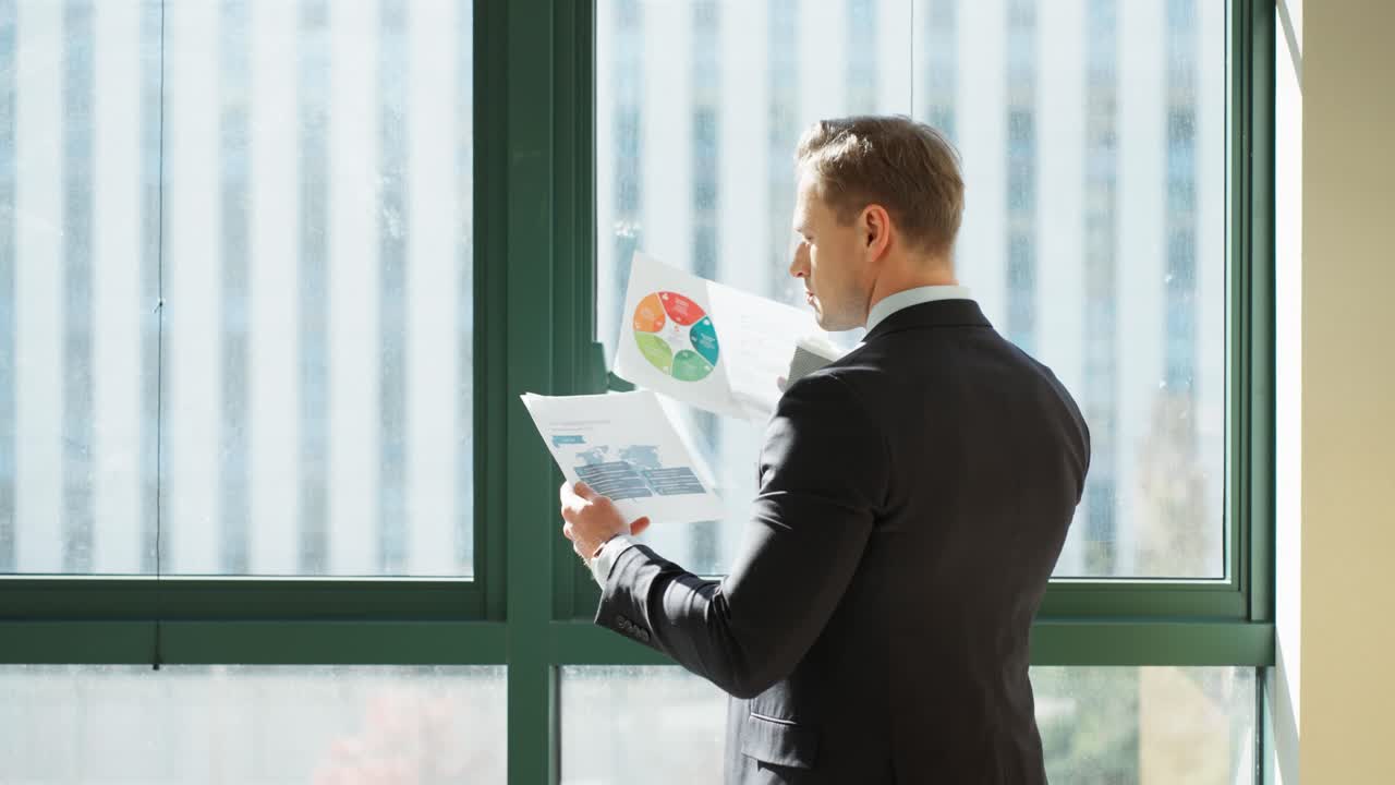 A businessman in a suit reviews documents with a colorful charts by office window, drinking coffee on camera dolly push-in