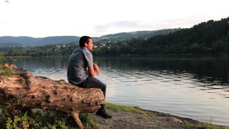 A-man-sits-alone-on-a-tree-stump-looking-out-over-a-lake-as-listens-to-music-wearing-ear-buds