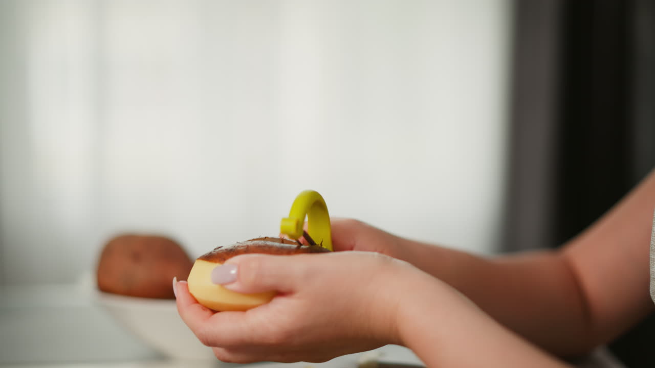Close up hand view of fair skinned woman peeling potato using green peeler beside bowl of sweet potatoes with soft blur kitchen background and natural light filtering through translucent curtain
