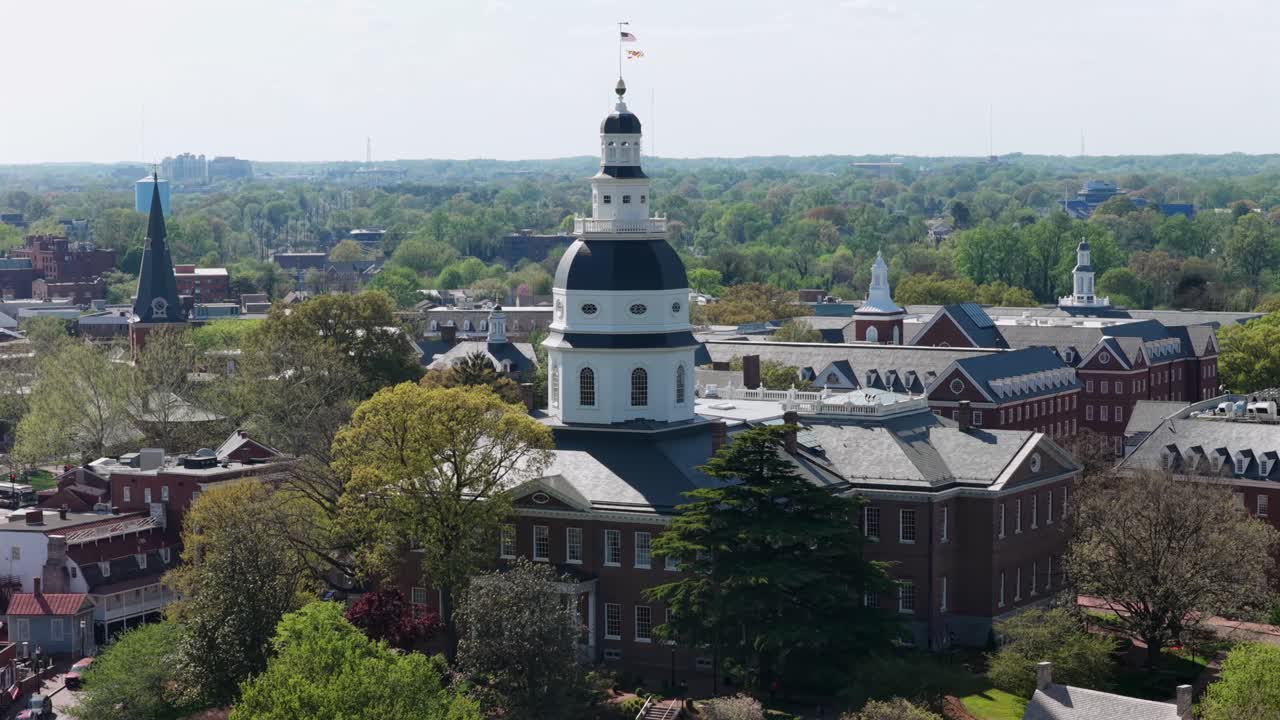 Luftfoto af tilgangen til Maryland State House med vajende amerikanske og Maryland-flag, omgivet af historiske bygninger og grønne træer under lyst forårssolskin i den naturskønne amerikanske hovedstad Annapolis.