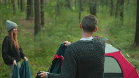 young man in green knitted sweater stands beside woman in gray beanie as they hold tent cover together, preparing to drape it over pitched tent in quiet forest surrounded by trees