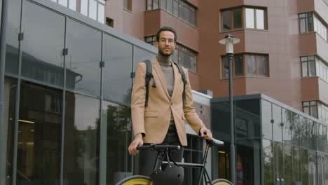 young american man in formal clothes and backpack standing in the street with his bike and looking at the camera