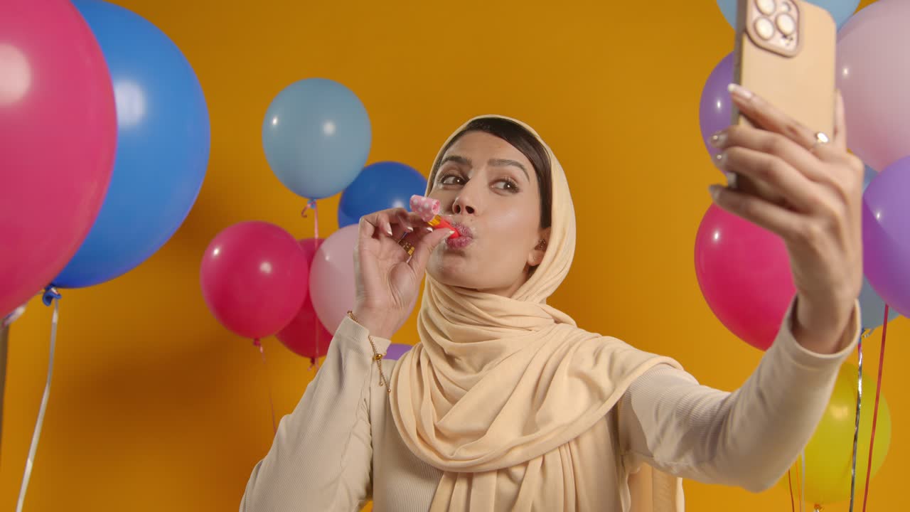 студийный портрет женщины, делающей селфи в хиджабе, празднующей день рождения с партийным вентилятором, окруженным воздушными шарами