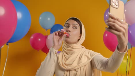 studio portrait of woman taking selfie wearing hijab celebrating birthday with party blower surrounded by balloons