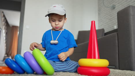 little toddler boy sitting on the floor collecting toy pyramid. baby boy wearing a cap playing at home. low angle view.