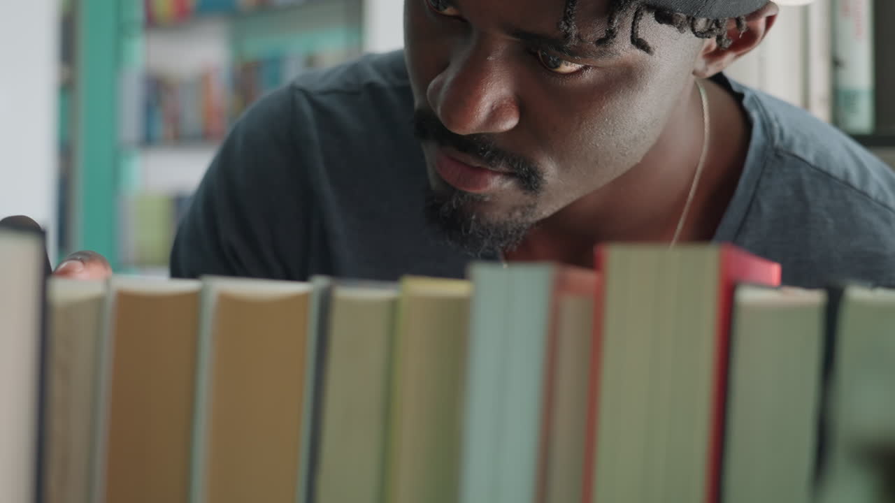 Close up of young man in casual shirt and backward cap leaning forward while browsing bookshelf in library, carefully scanning titles and pulling book slightly from row