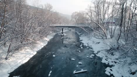 vista de pájaro del puente viejo, río congelado y árboles caídos cubiertos de nieve