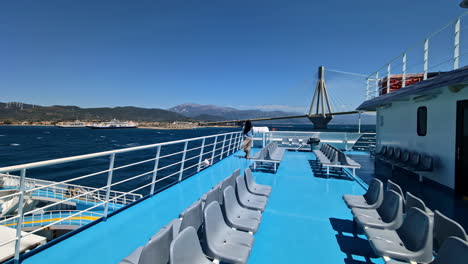 Young-woman-walking-on-windy-ferry-deck-with-Rio-bridge-in-background,-Greece