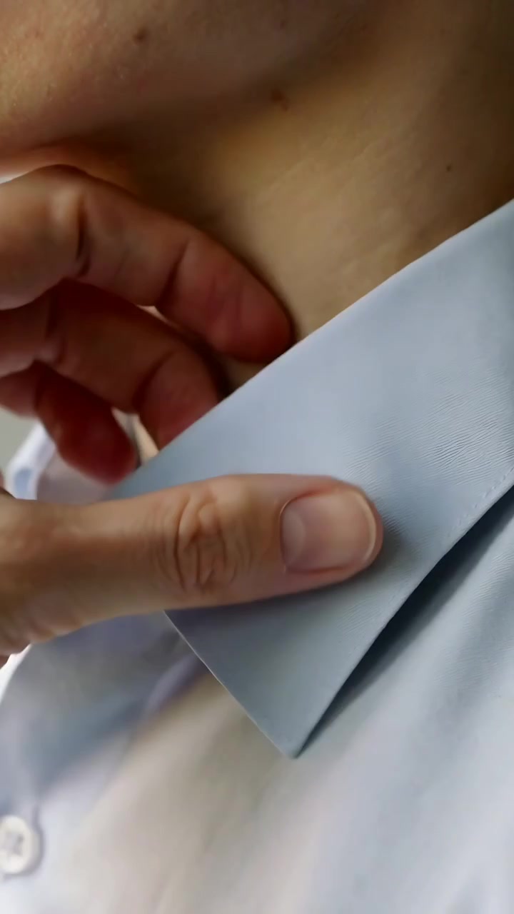 Video verticale: Una mano entra e sistema un colletto a punta su una camicia elegante chiara in una stanza, per una vestibilità perfetta.