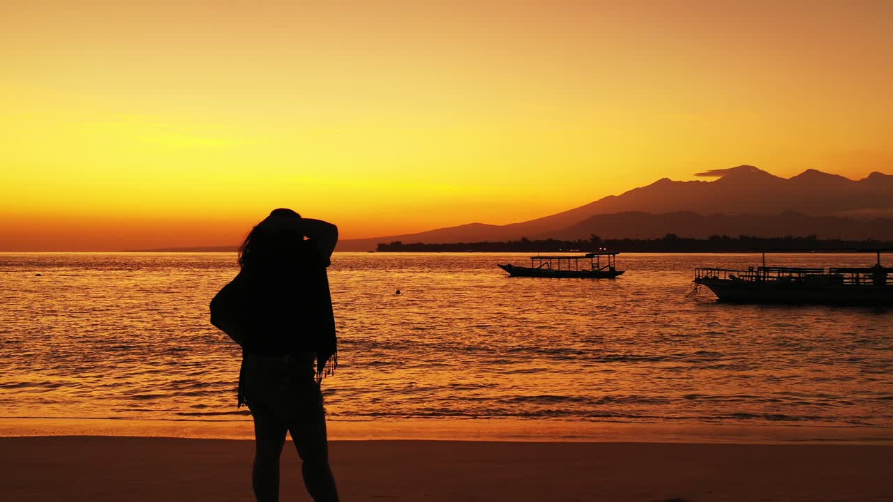 a woman standing on sandy beach during sunset in Hawaii with boats floating on the calm sea golden colored from the sun reflection