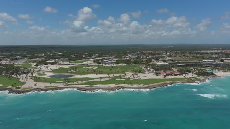 Broad aerial of golf resort along coastline showing greens, beach, and luxury waterfront homes, establishing panoramic drone