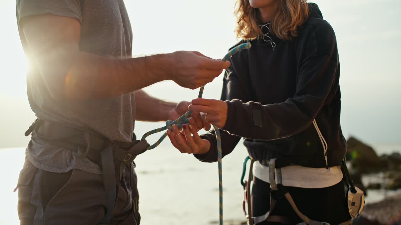 una foto en primer plano de una chica rubia con el cabello rizado en ropa negra ayudando a un tipo con una camiseta gris a atar un nudo especial en su fijación antes de comenzar la escalada en una orilla rocosa en un día soleado de verano