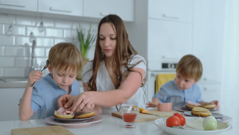 young mother with two young sons in the kitchen at the table preparing burger for lunch