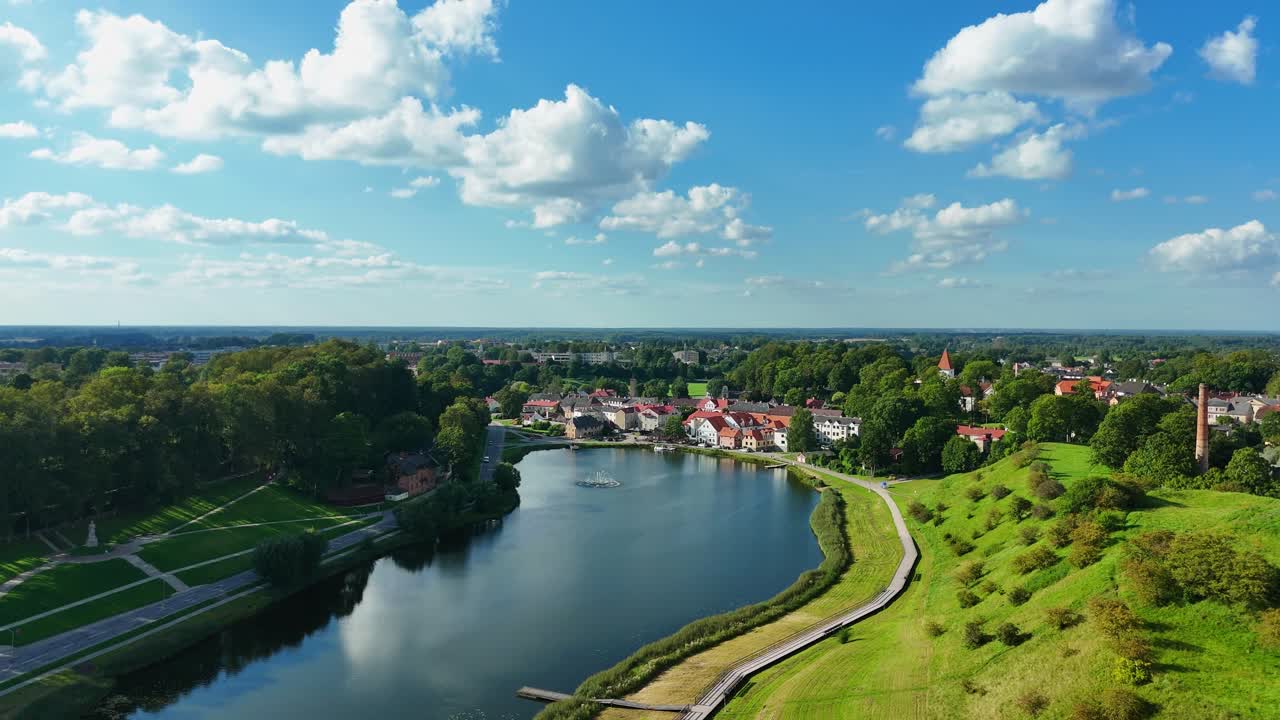 Aerial view capturing Latvian town Talsi, revealing historic center, mirrored lake surface, verdant parklands, and curving pathway under summer sunlight, drone pulling away