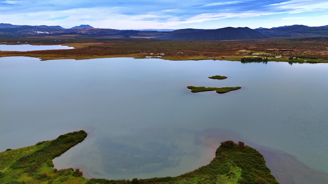Little curved island covered with grass is in the calm waterscape. Huge volcano crater at the backdrop of mountain range. Drone footage approaching the plain waterfront with poor vegetation. Iceland panorama from top.