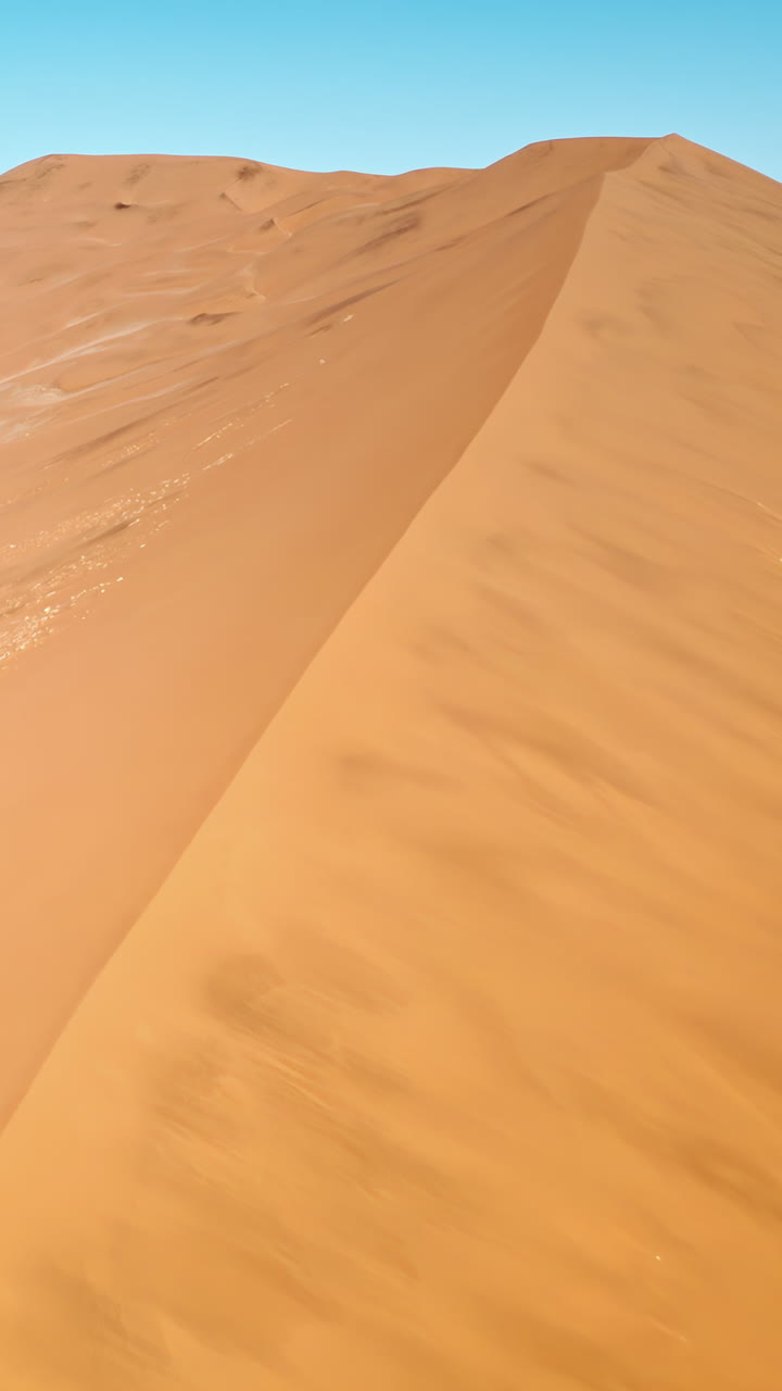 Vista aerea con drone lungo la cresta di una massiccia duna di sabbia nel deserto del Namib, con morbidi motivi modellati dal vento e un paesaggio desertico dorato. Verticale.