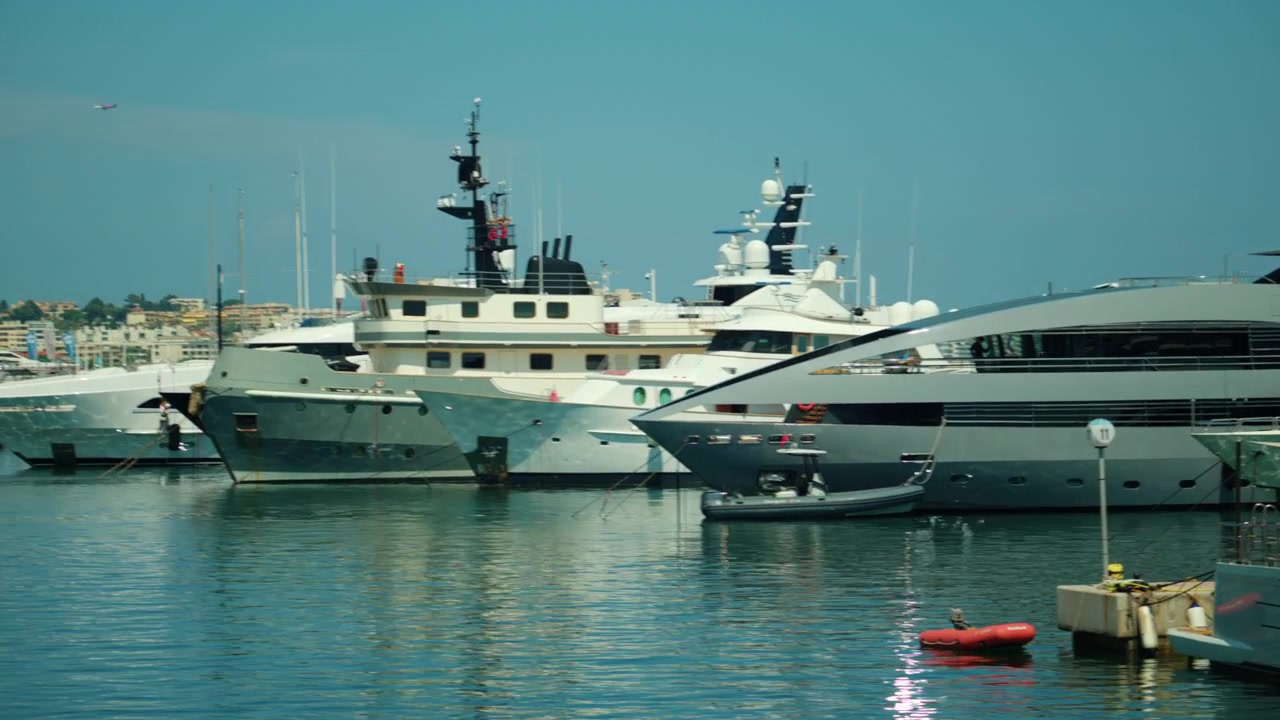 Molti yacht di lusso ormeggiati in un porto, che si riflettono sull'acqua calma con grandi edifici cittadini lungo la costa.