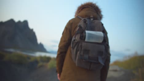 el excursionista se para con su mochila en una duna negra en la playa frente a un paisaje de montaña rocosa