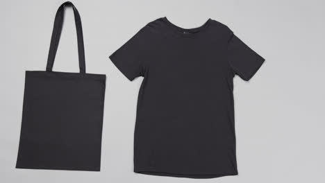 Primer-Plano-De-Bolsa-Negra-Y-Camiseta-Sobre-Fondo-Gris,-Con-Espacio-Para-Copiar,-Cámara-Lenta