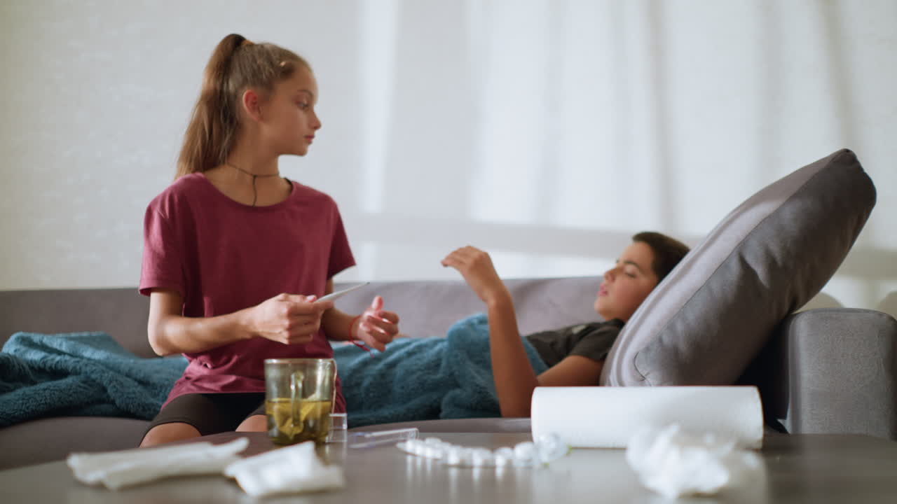 Young girl sitting next to sick brother, handing him thermometer to check his temperature, caring family moment, indoor setting with glass cup, tissues on table, sunlight through window