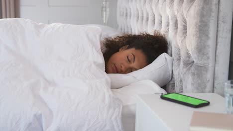 woman waking up in bed reaches out to turn off alarm on mobile phone