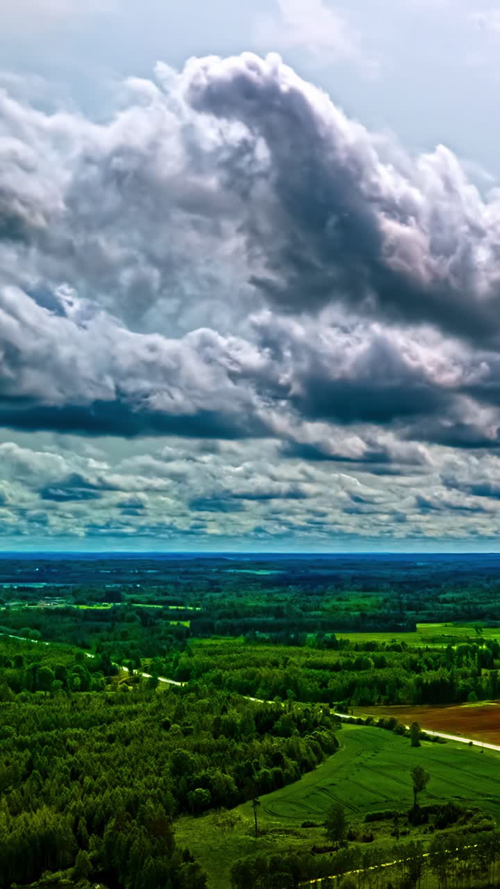 Hyperlapse aereo verticale in allontanamento che rivela la vasta campagna lettone con fitta foresta, verdi prati e campi sotto nuvole temporalesche scure che turbinano drammaticamente.