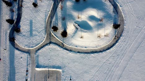 vista aérea de pájaro con vistas al invierno cubierto de nieve pista de patinaje ovalada de doble pez hecha por el hombre con una colina deslizante para que los niños vayan en trineo y camino de hielo enlaces a una pista de hockey rectangular pinos altos 1-3