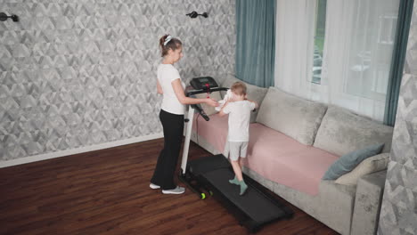 rear view of young boy wiping face with towel while exercising on treadmill at home, woman taking towel from him and hanging it back on treadmill as workout session continues in cozy living room