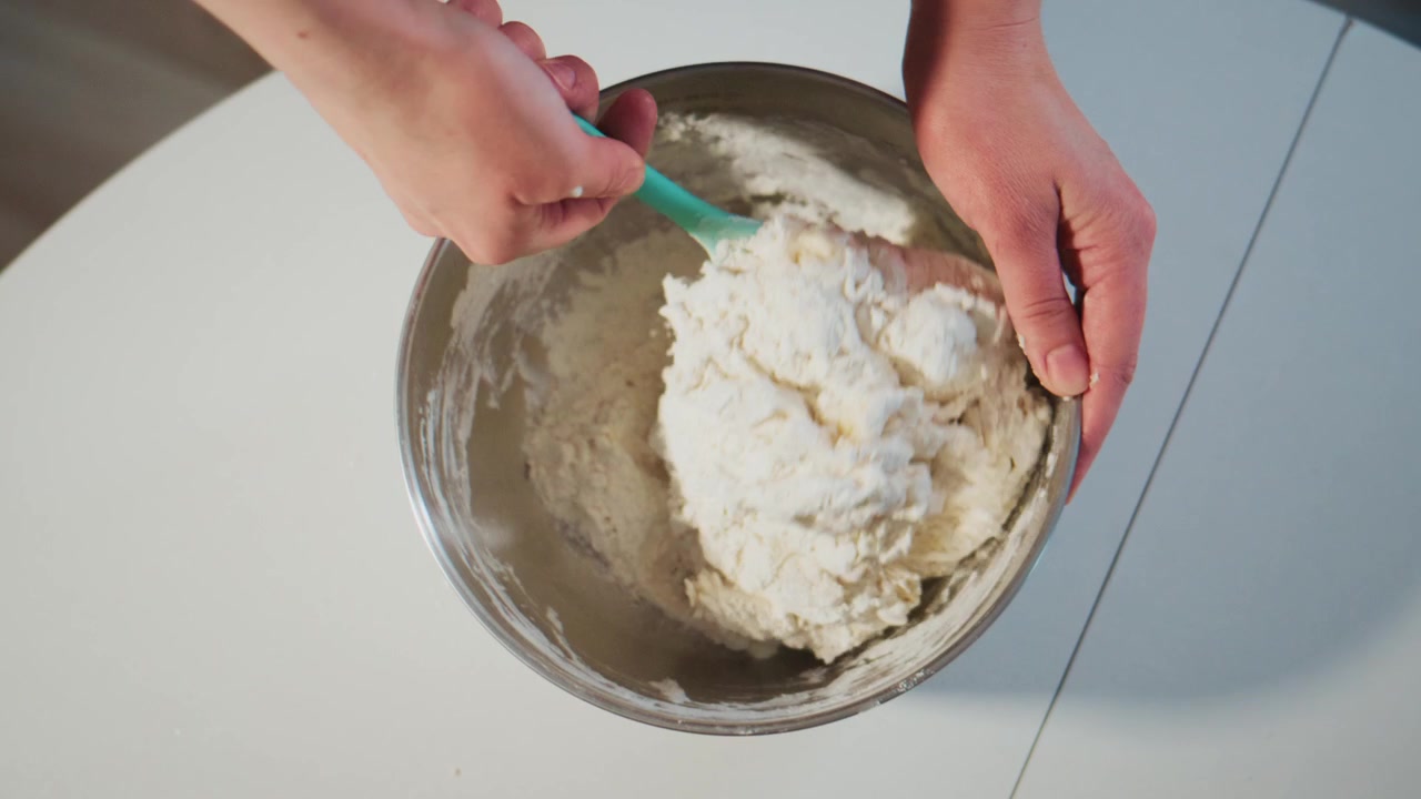 Hands mix thick dough in metal bowl. Close-up shows mixing action. White surface beneath. Ideal for baking tutorials or food blogs. Shows calm, focused kitchen activity