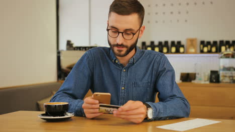 hombre hipster con anteojos usando tarjeta de crédito para comprar en línea con un teléfono inteligente mientras está sentado en un café