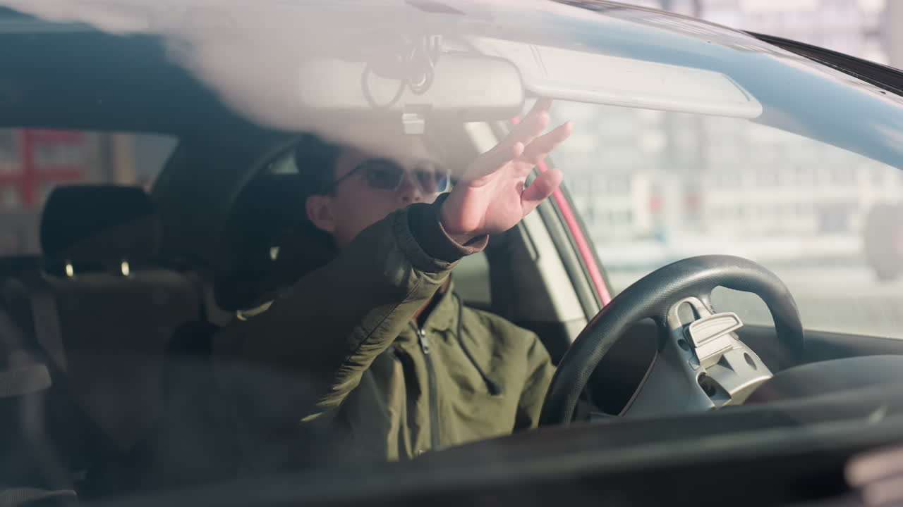 young boy in winter jacket seated in parked car lowers sun visor to block bright sunlight entering through windshield as urban buildings reflect in background on clear day