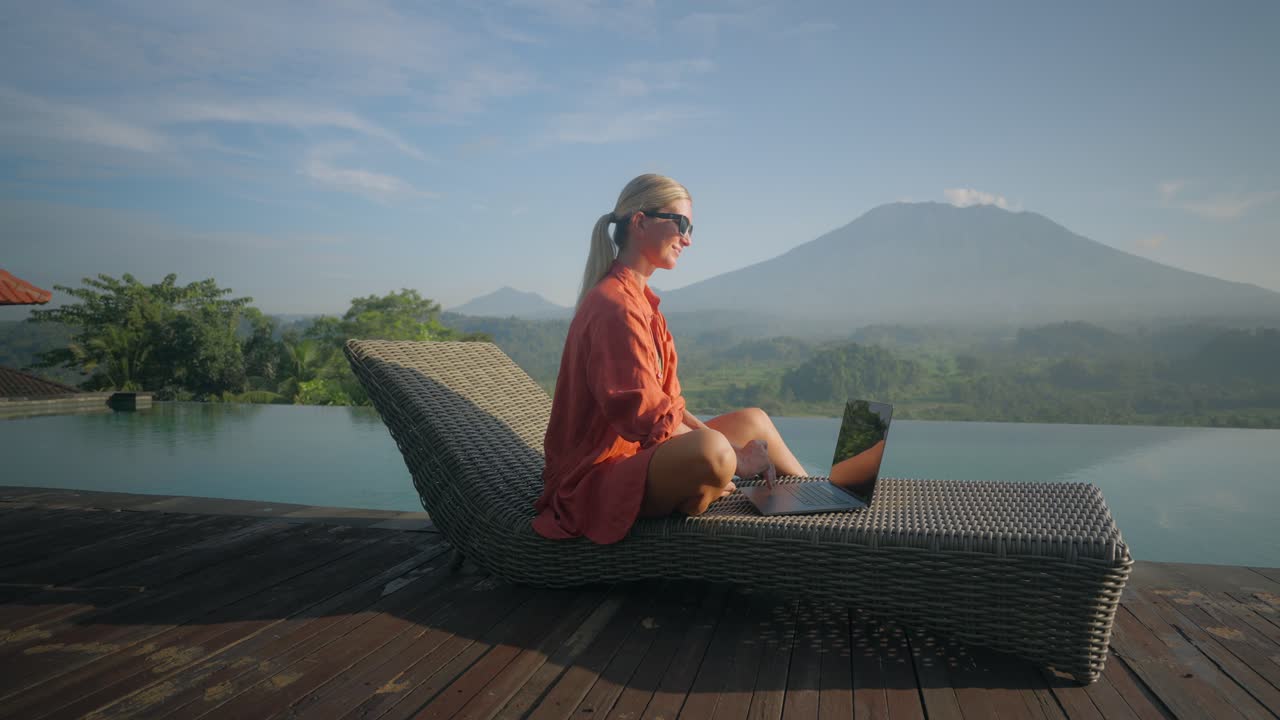 Hard working millennial woman typing on laptop laying backwards on lounge chair at infinity pool, incredible Bali landscape