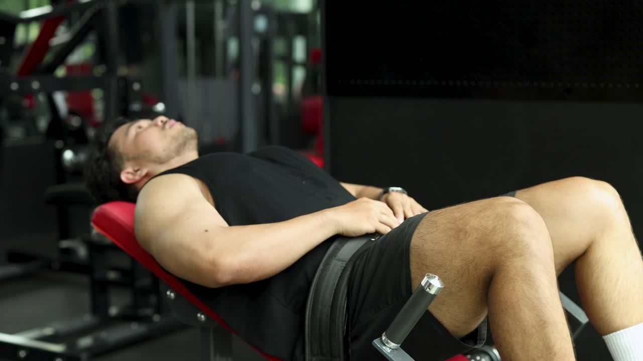 Male athlete performs hip thrust exercise on machine in modern, well-lit Bangkok fitness gym