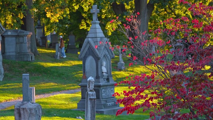 Una croce sovrasta un memoriale cimiteriale accanto a un fogliame rosso a Brooklyn. Pannelli di pietra appuntiti circondano una figura scolpita con erba e monumenti sparsi dietro l'arbusto