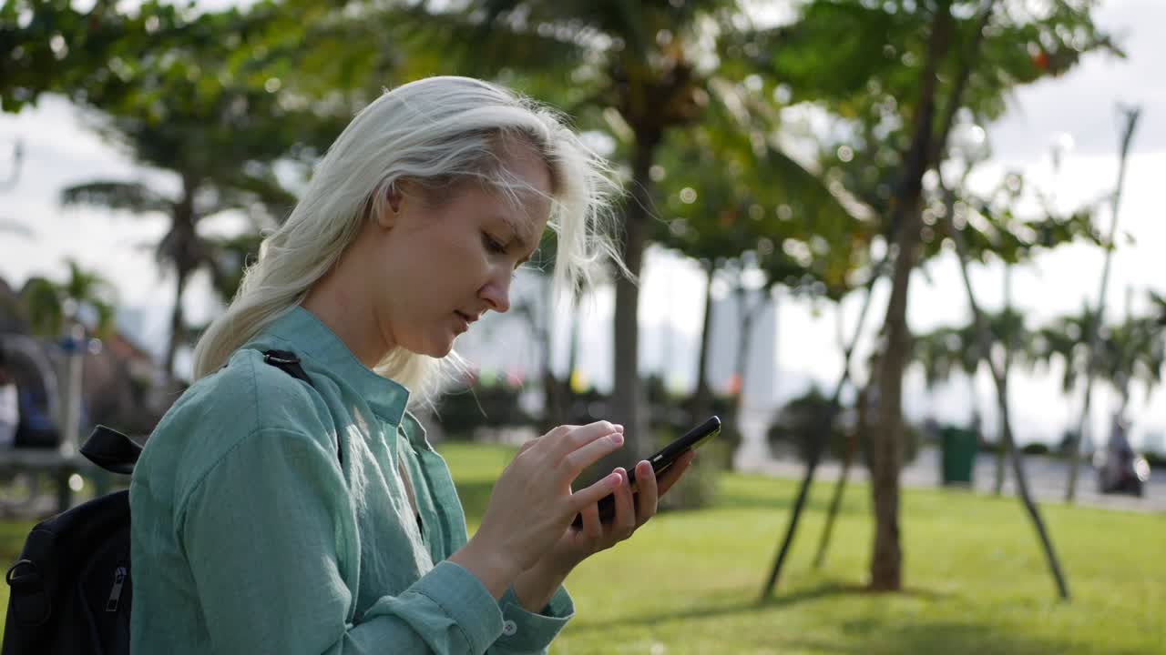 Beautiful slim woman with long blonde hair in green shirt sits on the ground and using smartphone over background the park. Girl on the square touching screen and smile