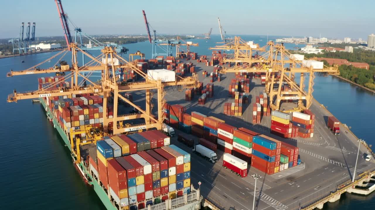 Gru a portale che movimentano un container su una nave verde alla banchina del porto, con camion che ricevono il carico.