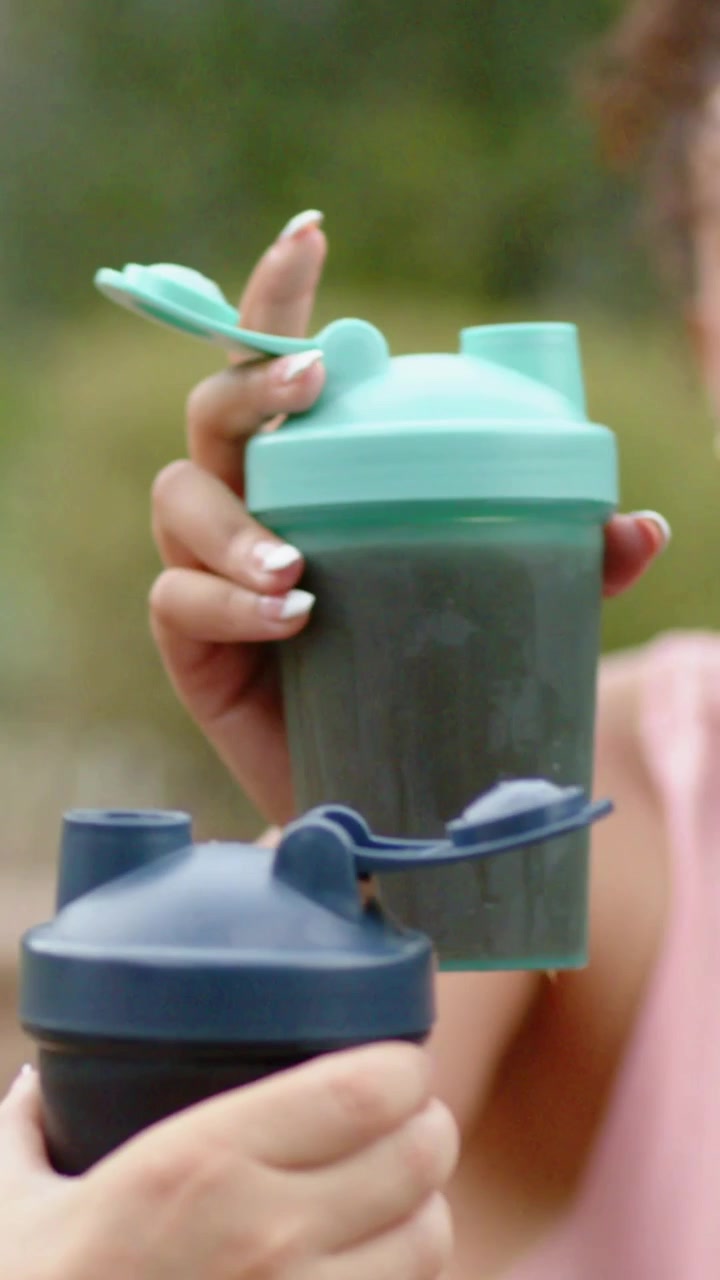 Video verticale: Donna che brinda con un top rosa, sollevando uno shaker color verde acqua al parco, con un sorriso che fa brillare uno shaker blu scuro.