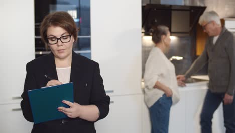 woman real estate agent filling in document on clipboard while aged couple examining kitchen on background