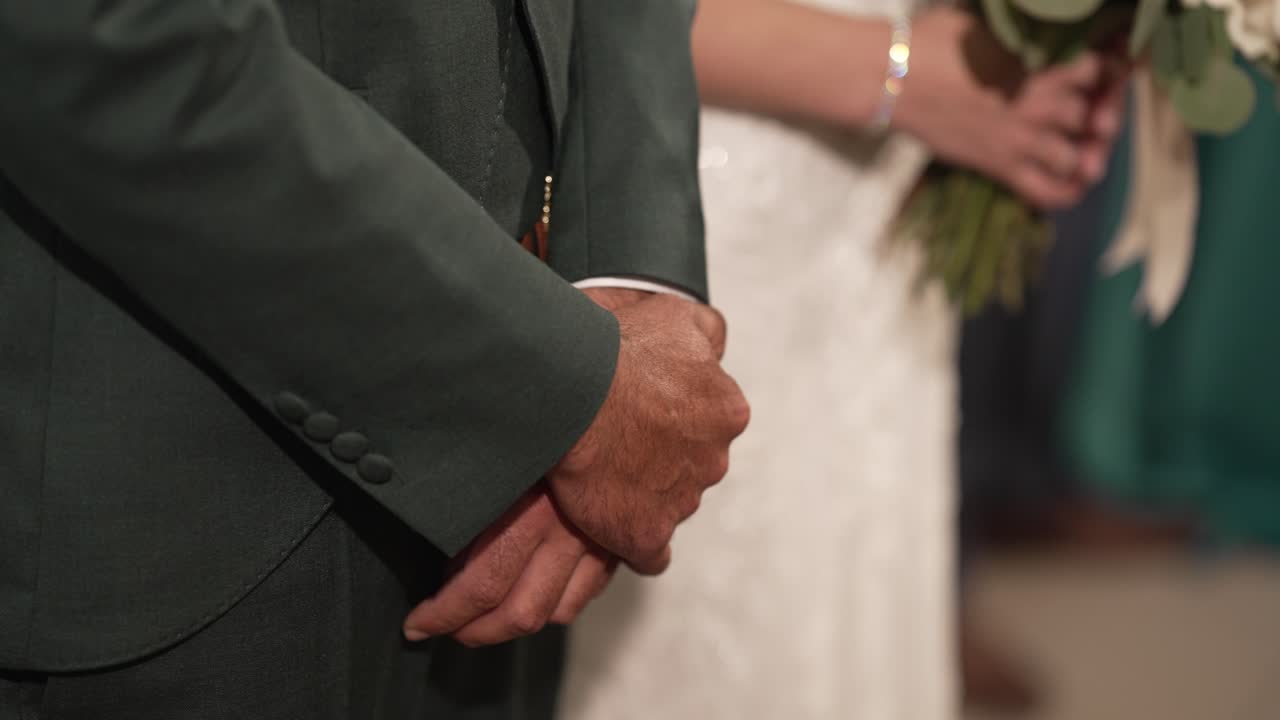 Close up of elderly man's hands clasped, bride holding bouquet in background