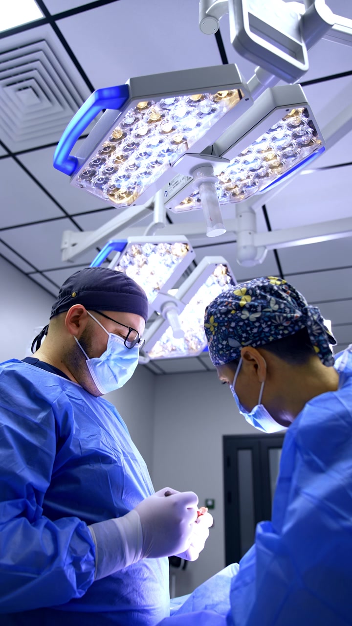 Dos médicos con uniformes azules están de pie bajo lámparas segmentadas en la sala de cirugía. Vista en contrapicado de los doctores realizando la operación. Video vertical.