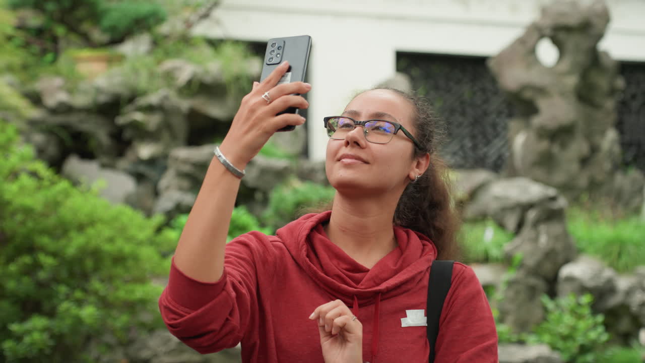 Woman holding smartphone in garden taking selfie, adjusting hair, smiling, checking photos, stone rockery backdrop, lush green bushes, casual red sweater, backpack strap visible, relaxed travel vibe