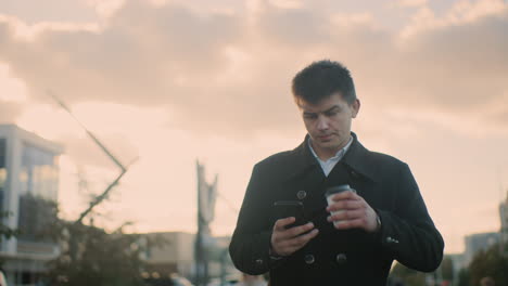 man walking outdoors, sipping coffee while browsing his phone with a contemplative expression in vibrant blur urban setting featuring modern buildings, parked cars, greenery, and people walking