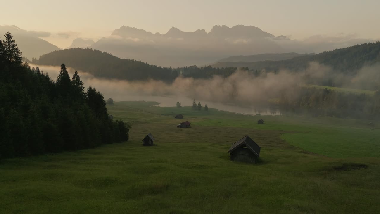 niebla matutina sobre wagenbrüchsee en alemania con montañas, campos verdes y cabañas de granja, vista aérea