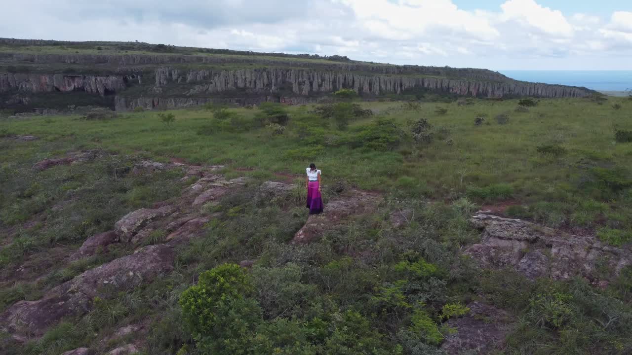 una mujer con un vestido colorido se agita ante la cámara en la cima de una dramática meseta de acantilados.